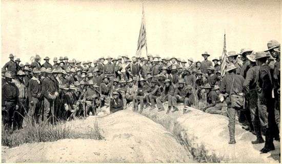 A black and white photo of US Army soldiers on 3 July 3, 1898, in an upside down V type formation on top of Kettle Hill; two American flags are center and right. Soldiers facing camera.