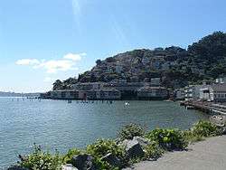 Cityscape containing a seafront and, mostly in the top right-hand corner, a hillside with houses. Shrubbery and asphalt are present in the foreground.