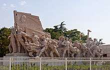 A brown stone sculpture of soldiers fighting with various weapons, led by one carrying a flag with a man's face in profile on it