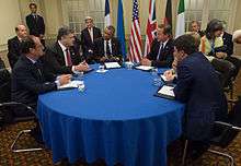 A group of men and women is Western business attire sit around a circular table with a blue tablecloth.