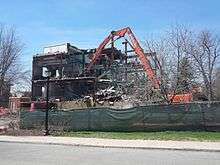 A clawed construction vehicle grabs at an exposed girder in a torn-open building.