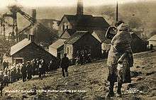 A young girl holds her younger sister on a hillside overlooking the Universal Colliery; a crowd, also waiting for news, are a little further down the hill.