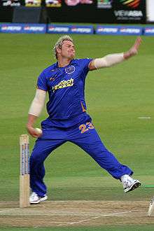 A white coloured man in his bowling action. He is wearing a blue outfit and is alongside cricket stumps. A field and boundary ropes can be seen in the background.