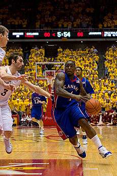 A person wearing a blue-colored basketball uniform dribbling a basketball running against another person wearing a white basketball uniform.