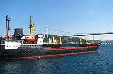 A pair of large ships sailing on a waterway, with a suspension bridge and hilly terrain in the background.