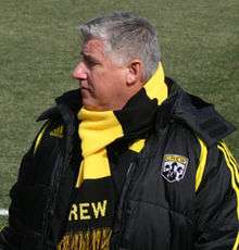 A portly man with graying hair. He is wearing a coat and scarf that are both black and yellow.