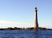 A tall lighthouse stands on low rocks under a blue sky.