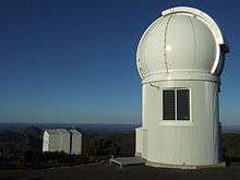 A dome atop a short cylinder sits on a rocky mountaintop. A square building sits further down the slope.