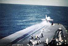 Colour photograph of a white military aircraft having just taken off from the flight deck of an aircraft carrier at sea