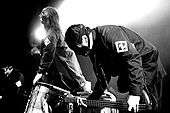 Black and white image of three men wearing jackets and masks over their faces. The one in the forefront is bend over, holding a guitar.