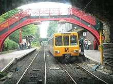 A metro train passing through a station with two platforms and tracks. In the foreground is a 19th-century pedestrian bridge.