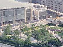 A large modern building of glass, metal and pale stone across the street from a garden with two small buildings.