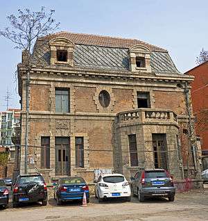 Side of the house with a stone bay window on the ground floor left, a tripartite French door on the right and an elliptical one in the middle of the upper story. Cars are parked in front.