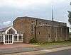 A bulky, plain brown brick building.  A flat-roofed projection on the near side has four evenly-spaced ground-to-roof windows with concrete details; the main part of the building behind it has six evenly-spaced windows in concrete surrounds, the leftmost of which is much taller than the others.  A tall, very thin green metal spire sits on the right side of the roof.  A modern porch with white cladding is attached to the left side of the building.