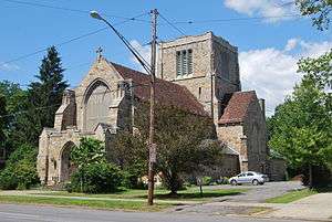 A brown stone building with a tall square tower in the rear and a pointed roof covered in red tile on a street corner with a street lamp in front