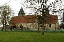 alt=A stone church with red tiled roofs seen from the south, showing the chancel, the south aisle and porch, and the tower with a pyramidal roof