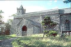 The nave of a stone church seen from the south with a porch and a west tower surmounted by a pyramidal roof