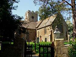 A view through a gate and graveyard to a yellow stone building with a square tower.