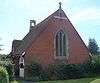 A red-brick church with a tiled roof extending nearly to ground level.  Low shrubs surround the building on all sides.  A three-light lancet window with stone mullions dominates the nearest side.  A small stone cross and bell-tower are on the roof.  The left-hand side has a white entrance porch.