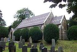 A small church with a decorated roof seen from the southeast. On the west gable is a simple bell tower.  In front of the church is a row of small yews and in the foreground are gravestones