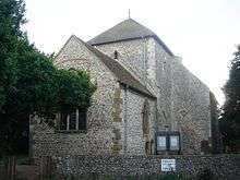 A flint rubble church with large stone quoins, behind a low flint wall.  A three-light lancet window in the nearest wall is partly obscured by a tree in full leaf.  A squat tower with a squared-off pyramidal roof stands in the middle.  To its right is a blank-walled protrusion with a blocked entrance.