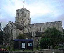 Side view of a substantial stone church with a square clock tower topped with a weather-vane.  The tower has two- and three-light arched openings in each face.  A triangular-roofed projection to the left has several round-arched windows and a small brown door.  The main body of the church, partly obscured by a tree, extends to the right, and has large buttresses topped with spirelets.