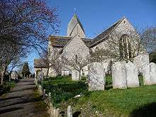 Side view of a flint church surrounded by a graveyard with bare trees and with a path to the left.  The top of a tower with a four-sided slate cap is visible behind the main body of the church.  A low porch protrudes to the left.