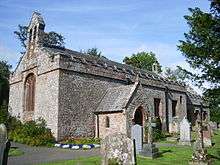 A small stone church seen from the southwest with two bellcotes and a battlemented parapet