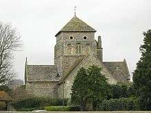 A cruciform church of flint and stone with a pyramid-roofed central tower topped with a weather-vane.  The nearest section is fully obscured by a tree.  The tower has three blocked round-arched windows and two round openings below the roof.