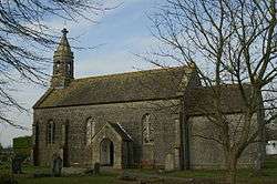 Small stone building with arched doorway. The church is partially obscured by trees.