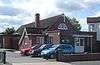 A long, low building of brick with a tiled roof, with extensions on the near side (next to a car park) and at the front. Behind the flat-roofed entrance porch is a semicircular window and a white crucifix.  The nearer of the extensions on the left has a small chimney-stack.