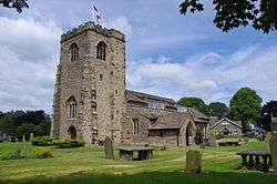 A stone church with a west tower, and a body of complex design including a porch and large rectangular paned windows in the clerestory.