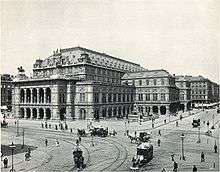  An imposing, heavily ornamented building in a city location, with numerous horsedrawn vehicles and pedestrians passing. There are visible tramlines in the street.