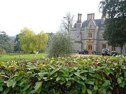 Stone manor house with tall chimneys. In front of the house are gardens and trees.