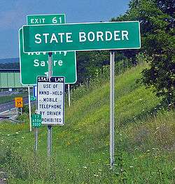 A white on green sign saying "State border". Behind it in the distance is a standard U.S. freeway exit sign.