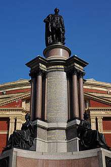 Photograph of an elaborate memorial centered on a massive stone column. At the base of the column there are two bronze statues of seated figures. There is a bronze statue of a man standing on top of the column. There are extensive carved inscriptions on the column itself. Behind the memorial is the brick and stonework facade of a large and elaborate building; there is a clear blue sky above the building.