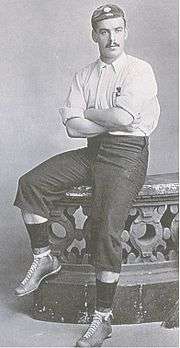 A man with a dark moustache, in a light-coloured football shirt, long dark shorts, and a cap, seated on the edge of a stone balustrade