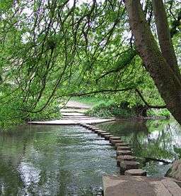 Wide river flowing from right to left. A series of seventeen hexagonal stepping stones lead across the river to the opposite bank in the distance.  Three wide low steps on the opposite bank leading from the river.            =