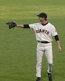 A man wearing a white baseball uniform, black hat and black glove stands in the field.