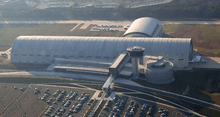 A large white building that resembles an airplane hangar. There is a large parking lot in the front with an enclosed glass bridge connecting it to the building.