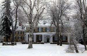 A snow-covered 2-story house with dormer windows on the roof and a porch on the ground floor. The stone house has several large trees in front.