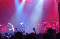 Two guitarists, a drummer, a bassist, and a vocalist are performing a song live on a stage lit by red and blue lights. The crowd, some of whom have their hands in the air, are visible in the foreground.