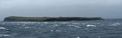 View of a low-lying island under grey skies, with rough water and cresting waves in the foreground