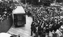 Crowds of people pack the area next to a road. Their attention is on a police van. Several of the watchers are pressing down on those in front of them or leaning over a wall. Police officers line the road.