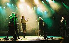 Four singers perform in the spotlights on stage. One is a female in shorts, a gauzy beige skirt and a leather belt, and plays the fiddle,  The males are wearing black sleeveless shirts, black pants and have military-looking gear. A rock band is in the background.