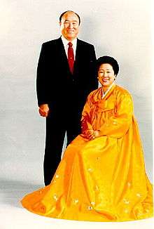 Moon (standing in suit) and his wife (seated in long yellow dress)