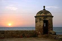 Old, somewhat decayed battlements, with a sunset in the background over the sea.