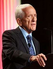 Waist high view of white haired man speaking at a microphone wearing a dark blue suit and tie