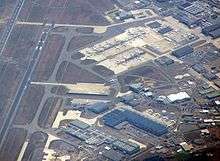 View from the air.  Runway to the left and bottom.  To the right long buildings and lots of aircraft.