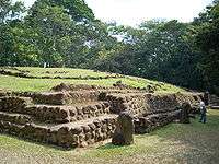 A low, wide structure with stepped sides, with the top covered in low grass. A dense treeline forms the backdrop to the structure. The right hand side of the structure has several standing stones before it, with a local guide standing by one of them.